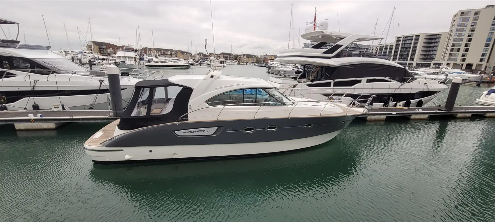 A sleek gray and white motorboat named "Flyer 12" docked at a marina surrounded by luxury yachts and buildings.